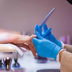 Stylistka w niebieskich rękawiczkach wykonuje przedłużanie paznokci na formie, precyzyjnie modelując płytkę.
